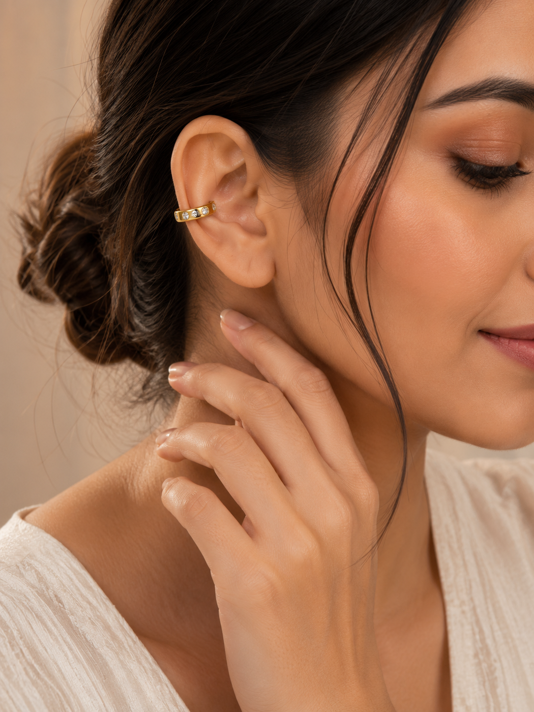 A close-up lifestyle shot of a woman wearing a delicate gold ear cuff with small clear stones, shown on her earlobe against a soft beige background.