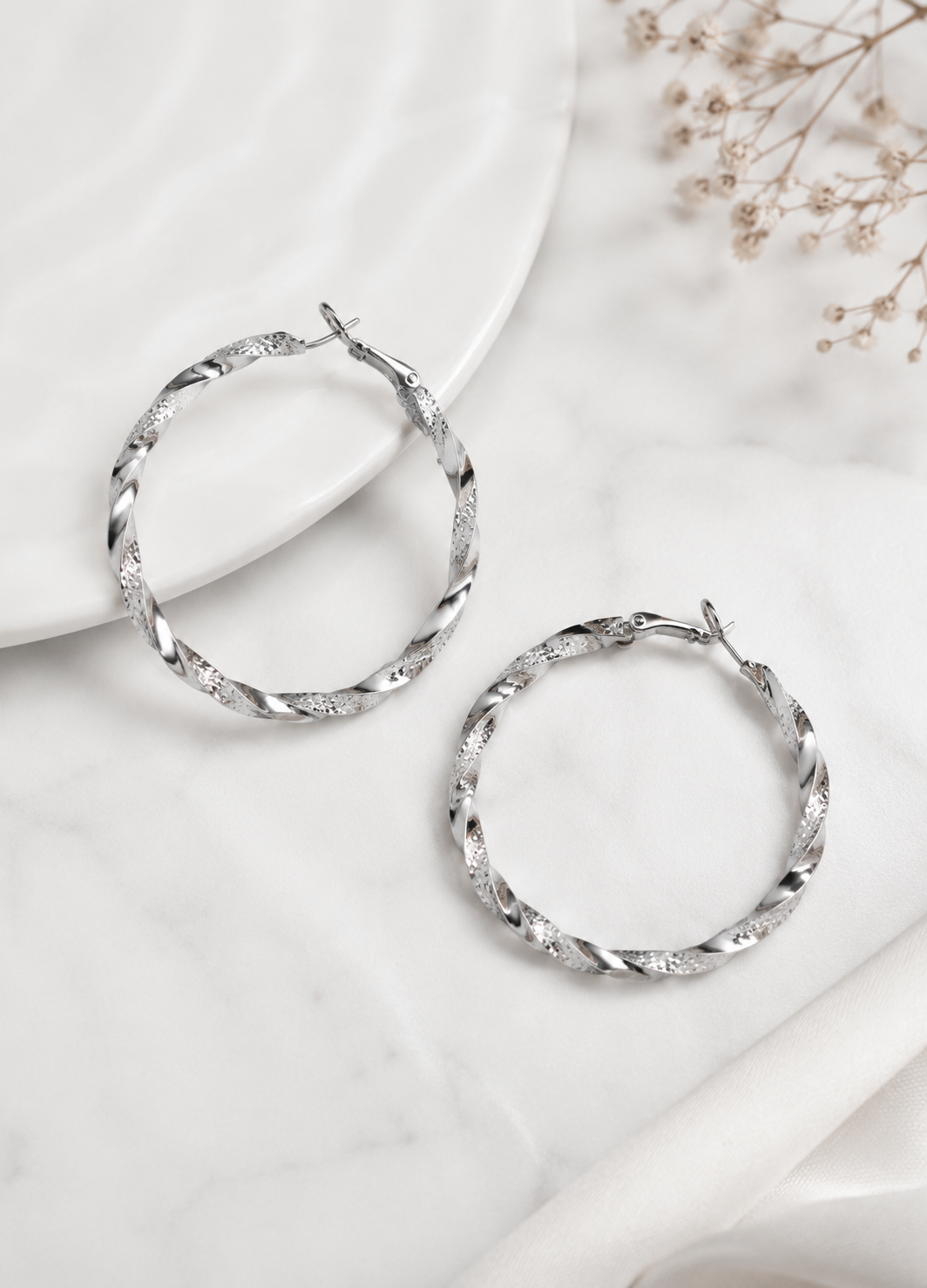 Pair of Orelva Celeste silver twisted hoop earrings displayed on a white marble surface.
